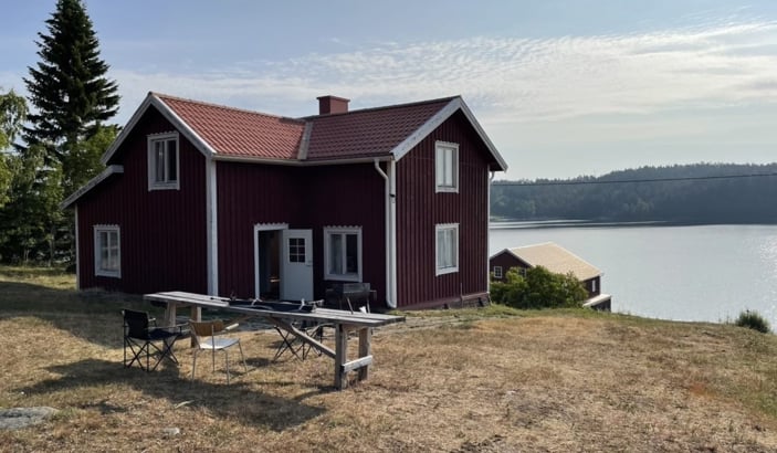 En röd stuga med vita knutar står på en höjd med utsikt över en sjö. Stugan har ett rött tegeltak och flera fönster, samt en öppen dörr. Framför huset finns ett enkelt utomhusbord med träbänkar och några stolar, placerade på en torr gräsmatta. I bakgrunden syns en mindre byggnad närmare vattnet, samt en skogsklädd horisont under en ljusblå himmel med tunna moln.