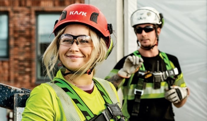 Två byggarbetare på en arbetsplats bär säkerhetsutrustning. I förgrunden syns en leende kvinna med en röd skyddshjälm märkt "KASK", skyddsglasögon och en neongrön arbetsjacka med reflexer. Hon har säkerhetsselar på sig. Bakom henne står en man i en vit skyddshjälm, solglasögon och en svart-grön arbetsjacka med reflexer. Bakgrunden visar en byggarbetsplats med en tegelbyggnad och en plasttäckning.