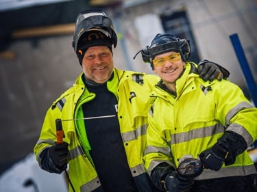 På bilden ser man två arbetskamrater posera. Den ena har svetsmask och den andra hörselkåpor. Båda har gula varseljackor på sig.